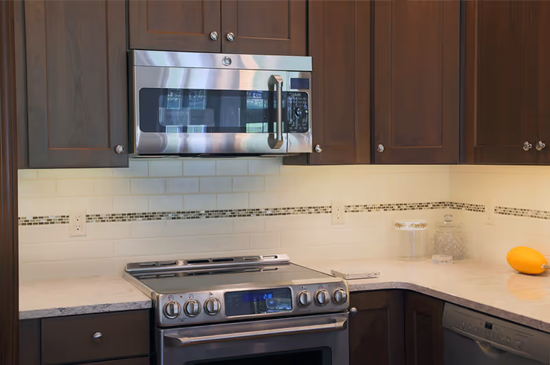 Modern kitchen with dark wooden cabinets, a stainless steel microwave mounted above a stainless steel electric stove, white subway tile backsplash with a decorative horizontal strip, and light-colored countertops with a few decorative items.