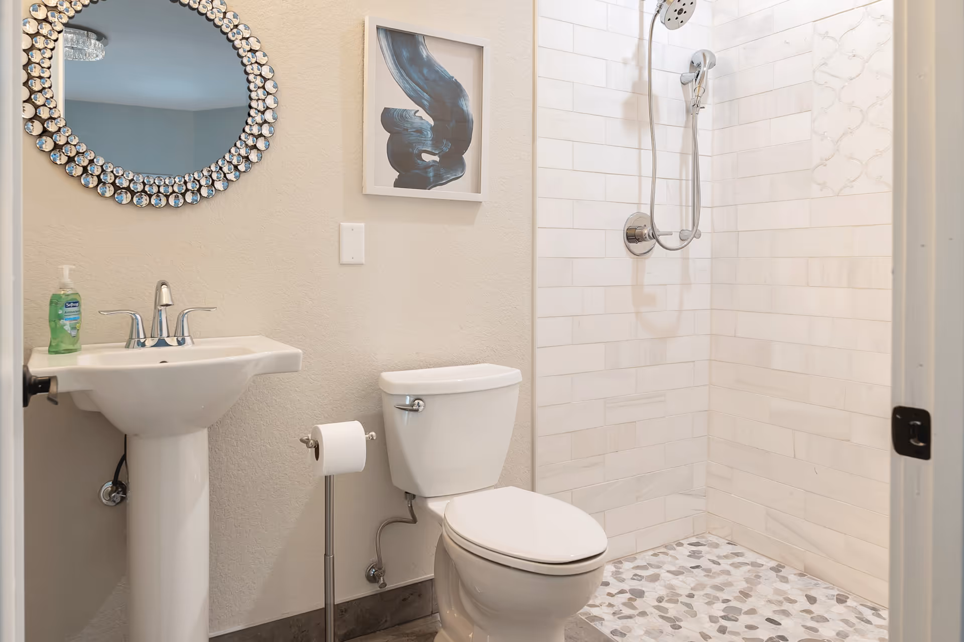 A clean and modern bathroom featuring a white pedestal sink with a chrome faucet, a decorative round mirror with a beaded frame, a framed abstract artwork on the wall, a white toilet with a standing toilet paper holder, and a walk-in shower with white subway tiles and a pebble stone floor.