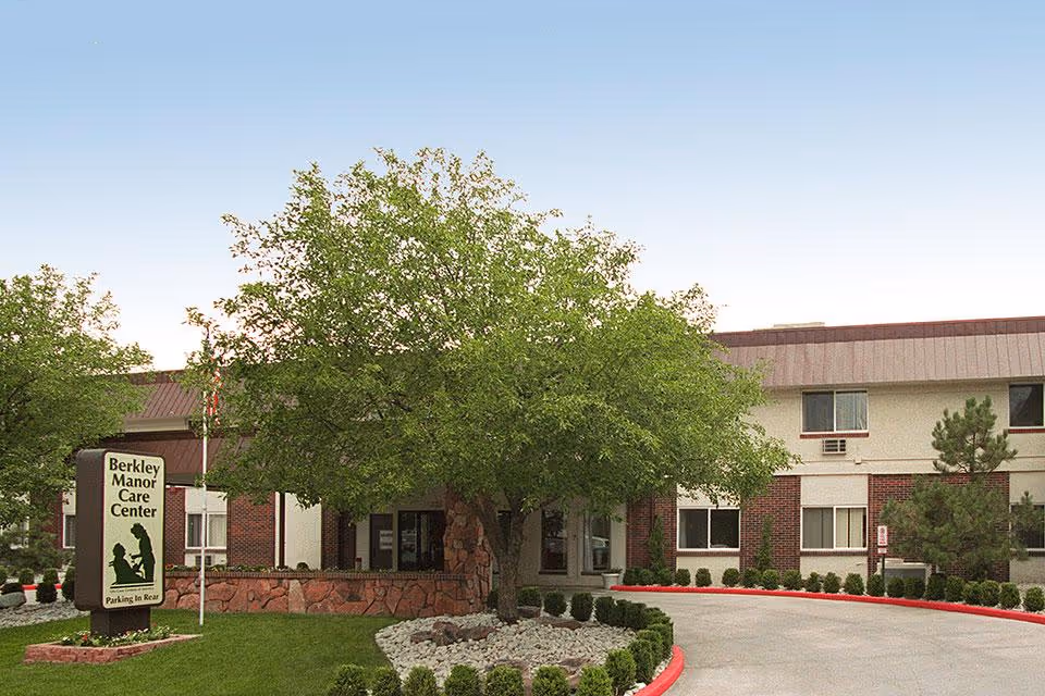 Exterior view of Berkley Manor Care Center showing a two-story building with a brick and light-colored facade, a large tree in front, a curved driveway, and a sign indicating the facility name and parking in the rear.