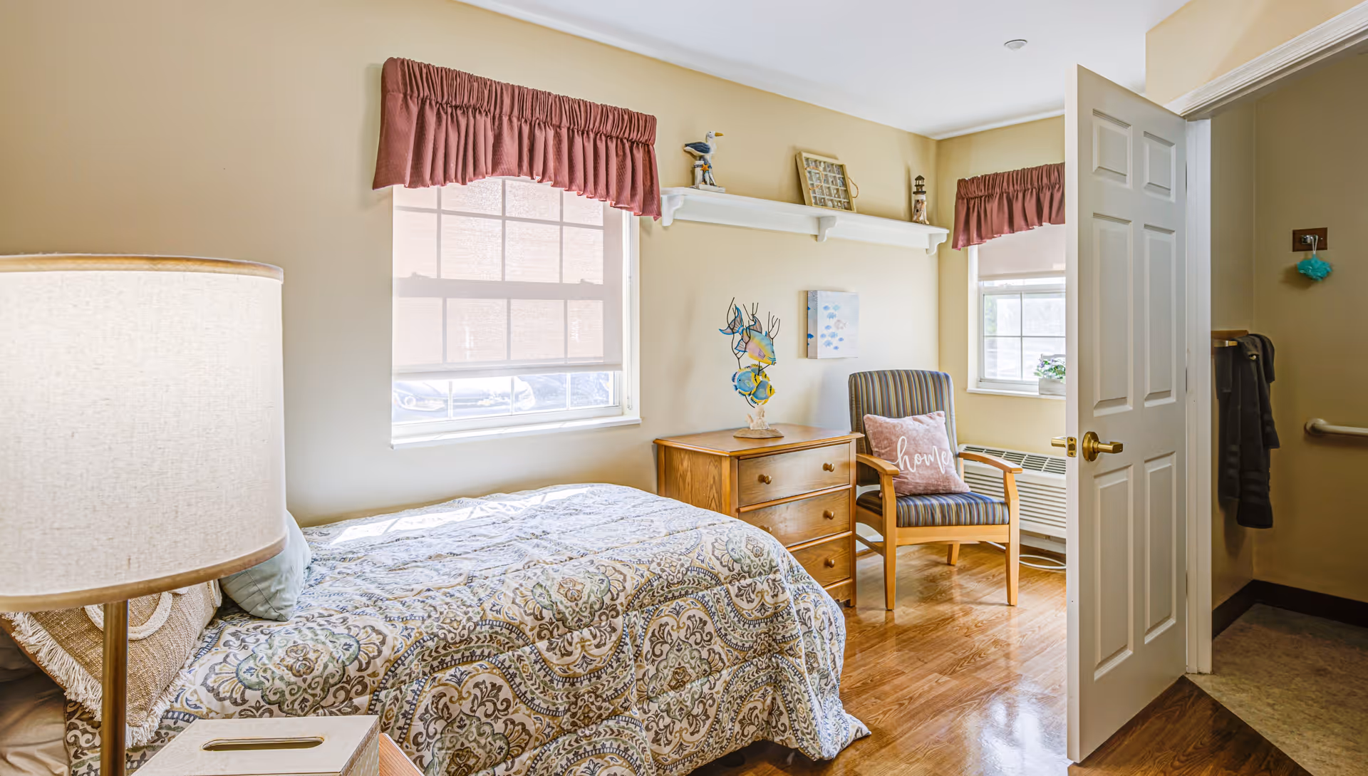 A cozy bedroom with a single bed covered in a patterned quilt, a wooden nightstand with a lamp, a wooden dresser with decorative items, a striped armchair with a pink pillow that says 'home', two windows with pink valances, and an open door leading to another room.