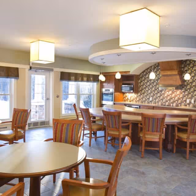 A bright and spacious kitchen and dining area with multiple wooden chairs around round and rectangular tables. The kitchen features modern appliances, a tiled backsplash, pendant lights, and a large island with seating. Large windows and a glass door allow natural light to fill the room.