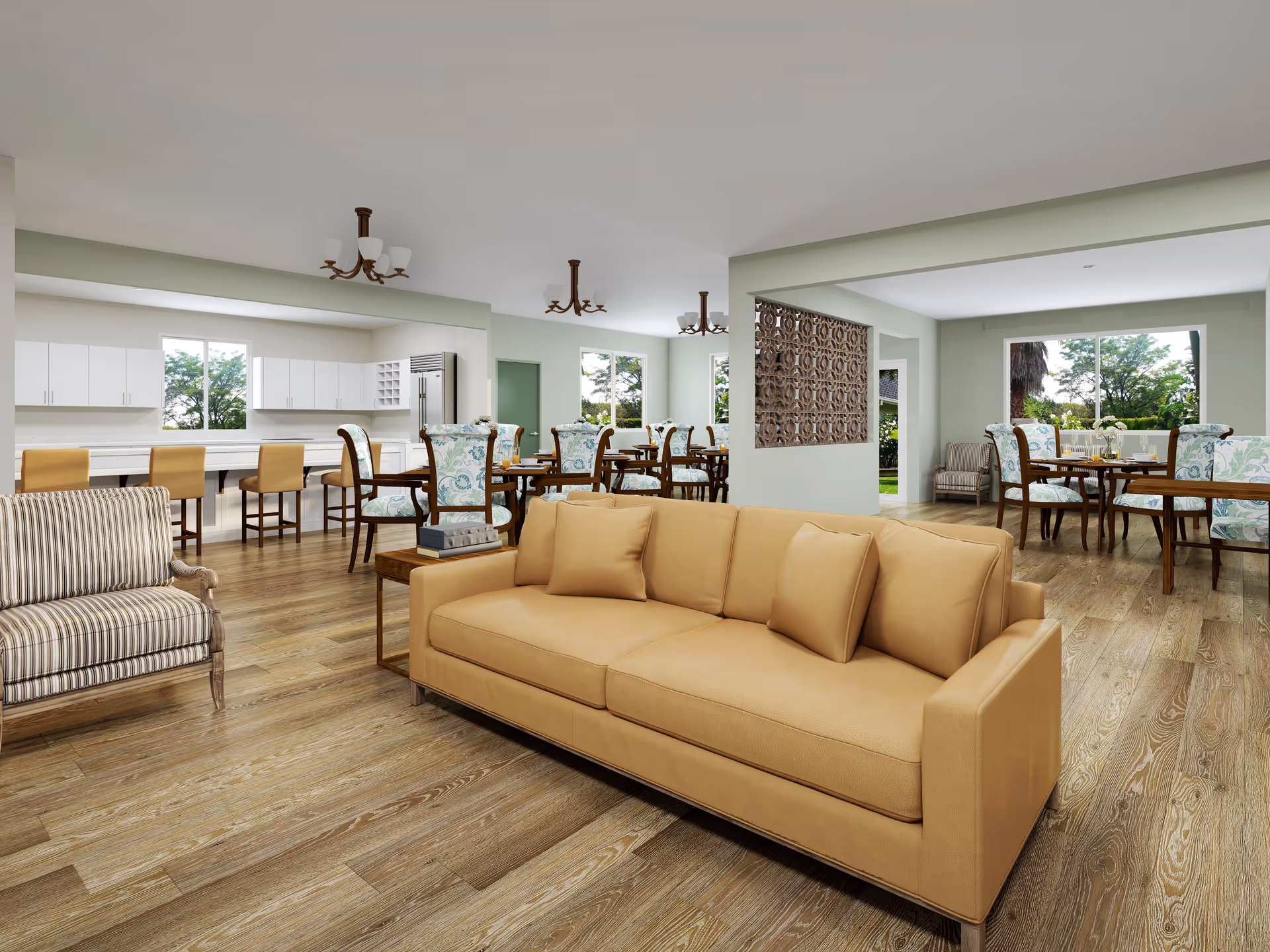 A spacious senior living common area featuring a tan leather sofa and a striped armchair in the foreground. Behind the seating area, there are multiple dining tables with floral upholstered chairs. The room has large windows letting in natural light and a kitchen area with white cabinets and bar stools in the background. The flooring is wood, and the walls are painted light green and white.