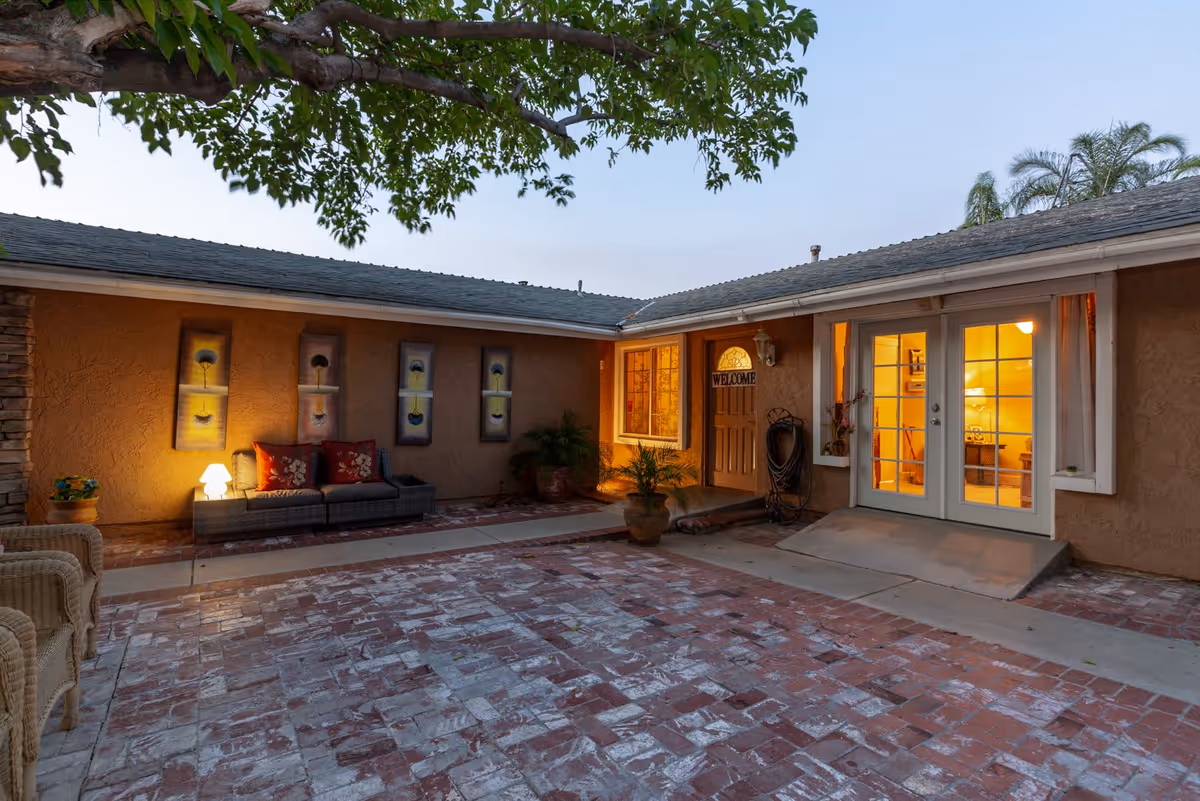 Outdoor courtyard area of a senior living facility at dusk with brick flooring, a wicker chair, a cushioned bench with pillows, potted plants, wall art, and a warmly lit entrance door with a welcome sign and glass double doors.