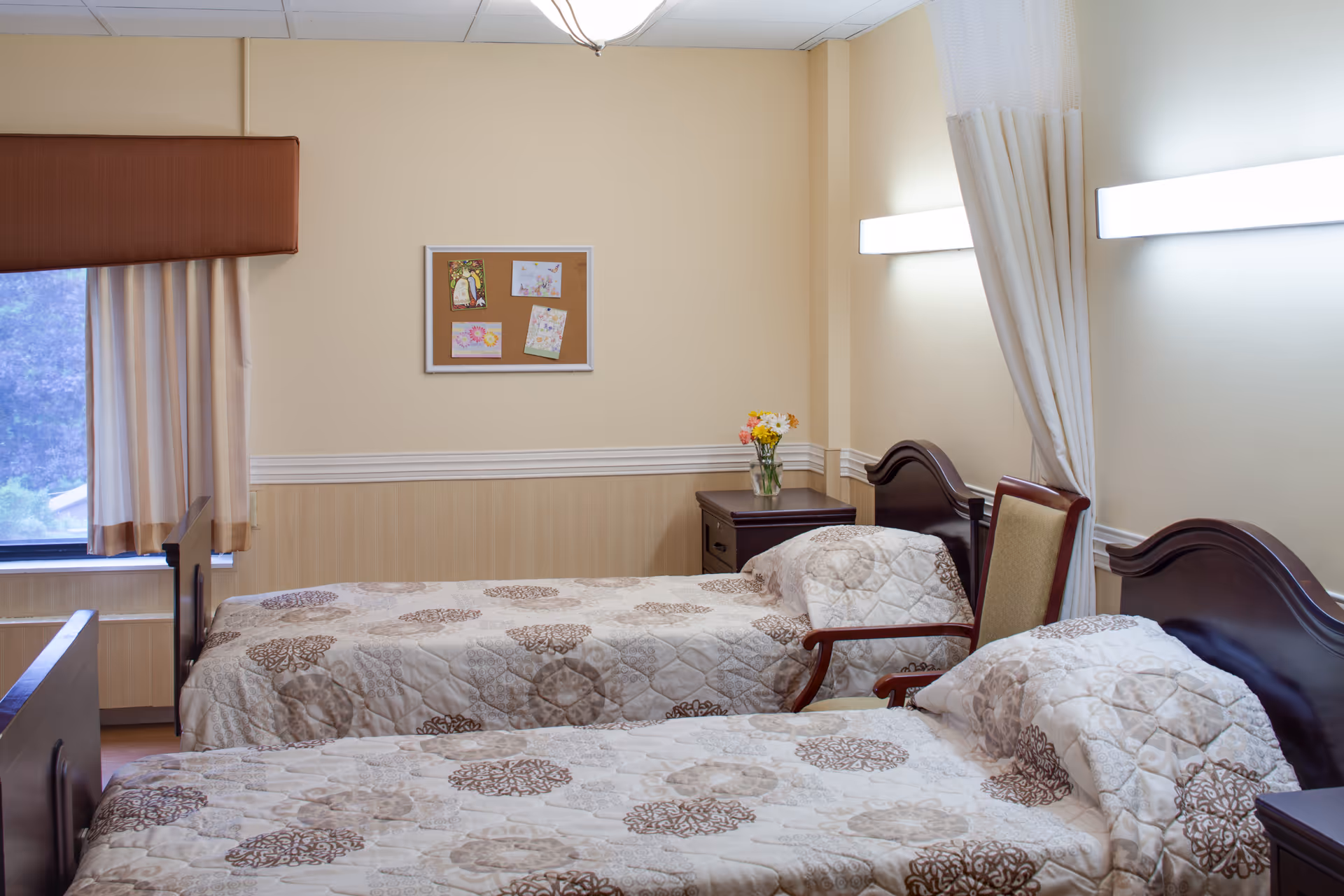 A cozy bedroom in a senior living facility with two beds, a window with curtains, and a bulletin board.