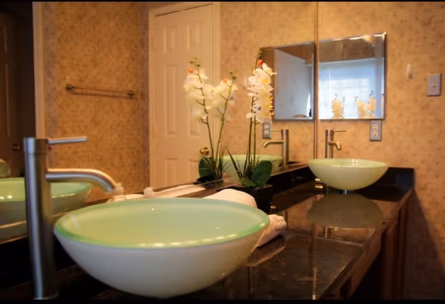 A bathroom countertop with two green glass vessel sinks and modern faucets. There is a large mirror above the countertop reflecting the sinks and a white door in the background. A white orchid plant in a black pot is placed between the sinks. The walls have a light floral wallpaper.