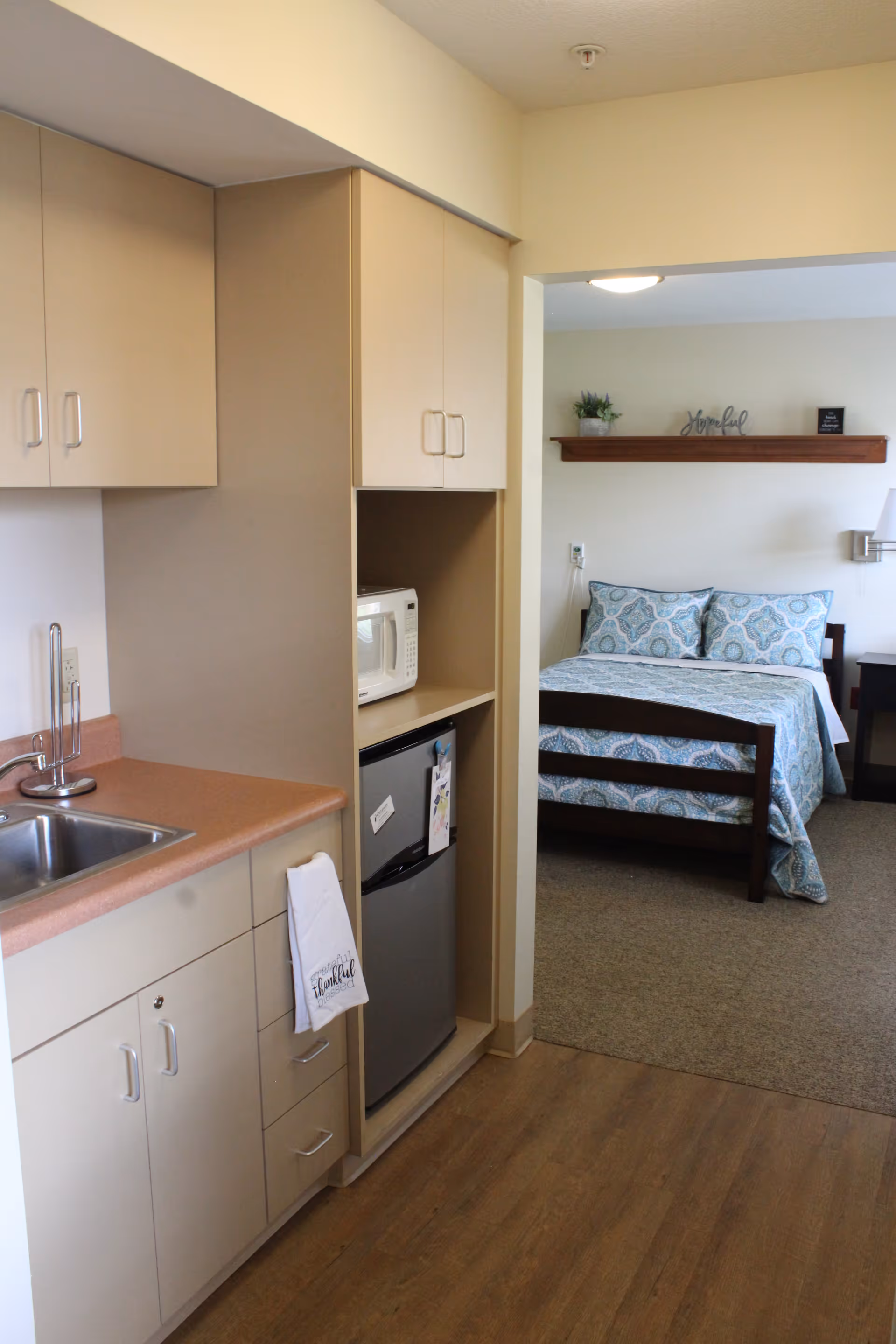 View of a small kitchenette area with a sink, countertop, cabinets, a microwave, and a mini refrigerator. Beyond the kitchenette, there is a bedroom with a bed made with blue patterned bedding, a wooden headboard, a nightstand, and a wall shelf with decorative items.