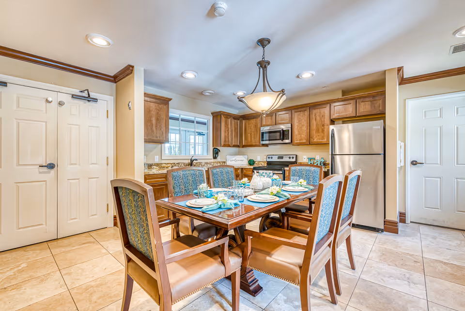 Dining table set for six in a bright kitchen/dining area with wooden cabinets and stainless steel appliances.