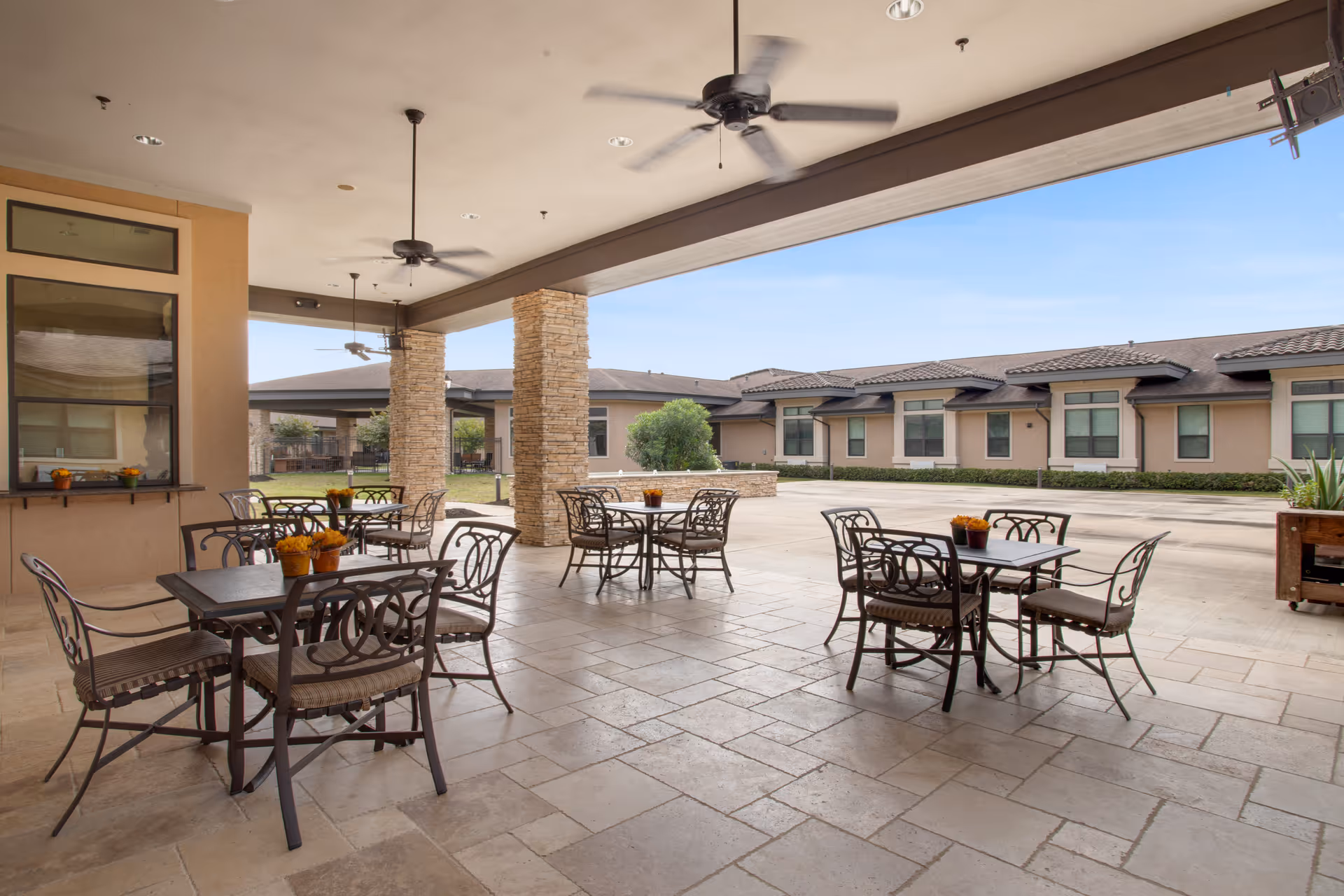 Covered outdoor patio area with several metal tables and chairs arranged for seating. The patio has ceiling fans and stone pillars, overlooking a courtyard with a building in the background under a clear blue sky.