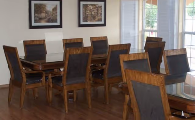 Wooden dining table with multiple matching chairs in a bright room with framed pictures and large windows.