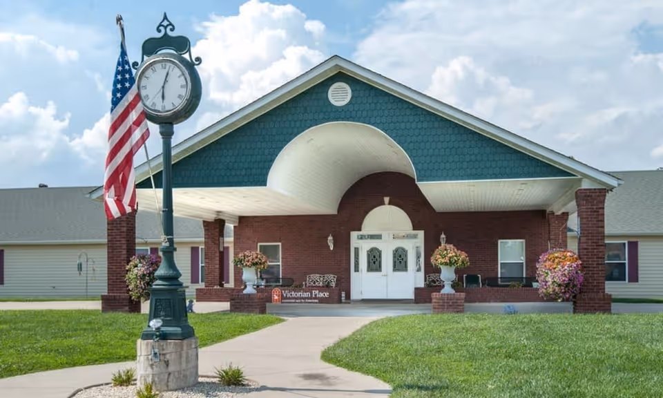 Front exterior view of Victorian Place of St. Clair, featuring a covered entrance with brick pillars, white double doors, decorative flower pots, and a vintage-style clock with an American flag beside it.