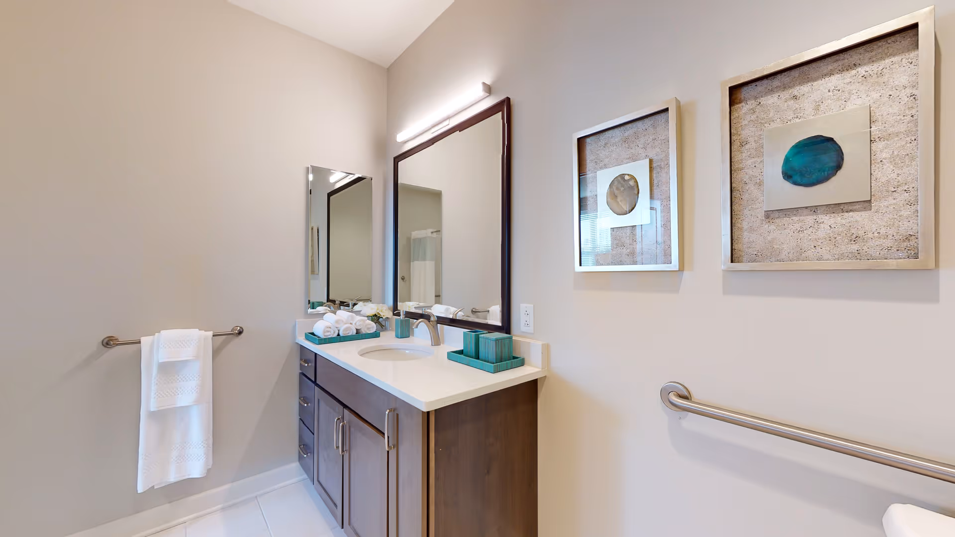 A clean and modern bathroom featuring a white countertop with a sink, dark wood cabinetry, a large mirror above the sink, and a smaller mirror on the adjacent wall. There are two framed abstract artworks on the wall, a towel rack with a white towel, and a grab bar for accessibility.
