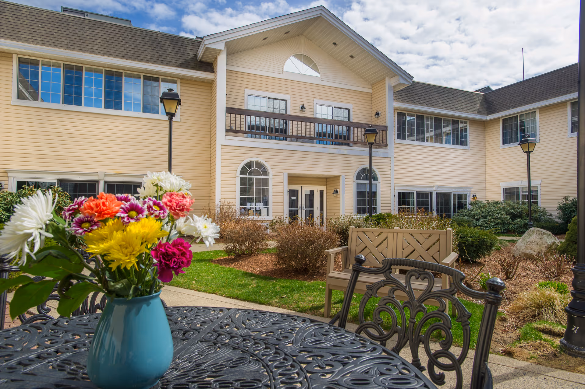 Outdoor courtyard area of a senior living facility with a black metal table and chairs in the foreground, a blue vase with colorful flowers on the table, beige two-story building with large windows and a balcony in the background, green grass, bushes, and lamp posts under a partly cloudy sky.