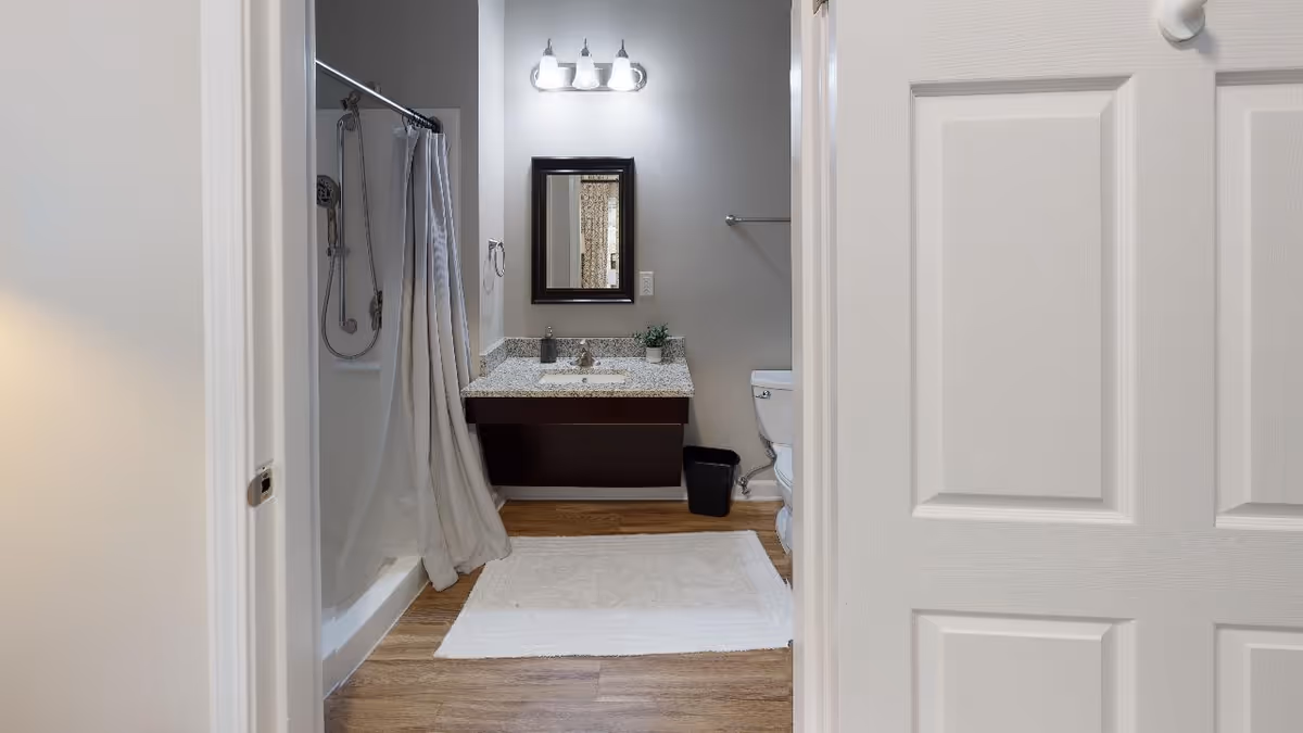 View into a bathroom with a white door partially open. Inside, there is a shower with a curtain on the left, a vanity with a granite countertop and a mirror above it in the center, and a toilet on the right. The floor is wood-style with a white bath mat in front of the vanity.