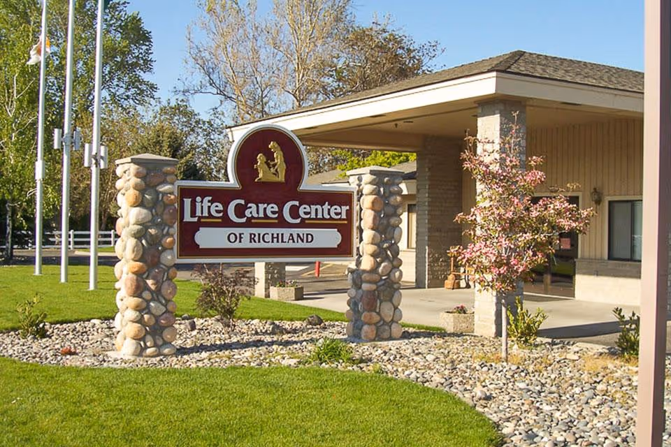 Outdoor view of the entrance to Life Care Center of Richland featuring a stone pillar sign with the facility name, a covered drop-off area, green lawn, small trees, and a clear blue sky.
