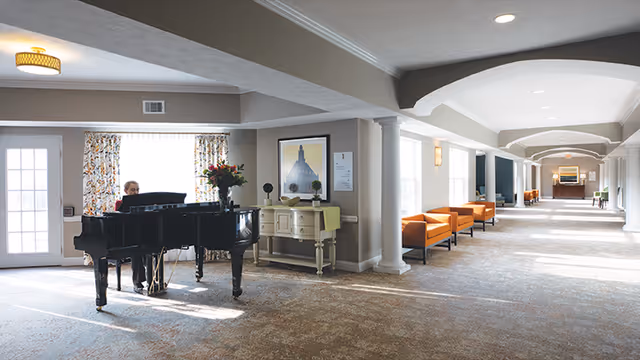 A spacious, well-lit common area in a senior living facility featuring a black grand piano with a person playing it. The room has large windows with patterned curtains, a side table with flowers and decorative items, and a row of orange chairs along the wall. The area has a carpeted floor, white columns, and a high ceiling with recessed lighting.