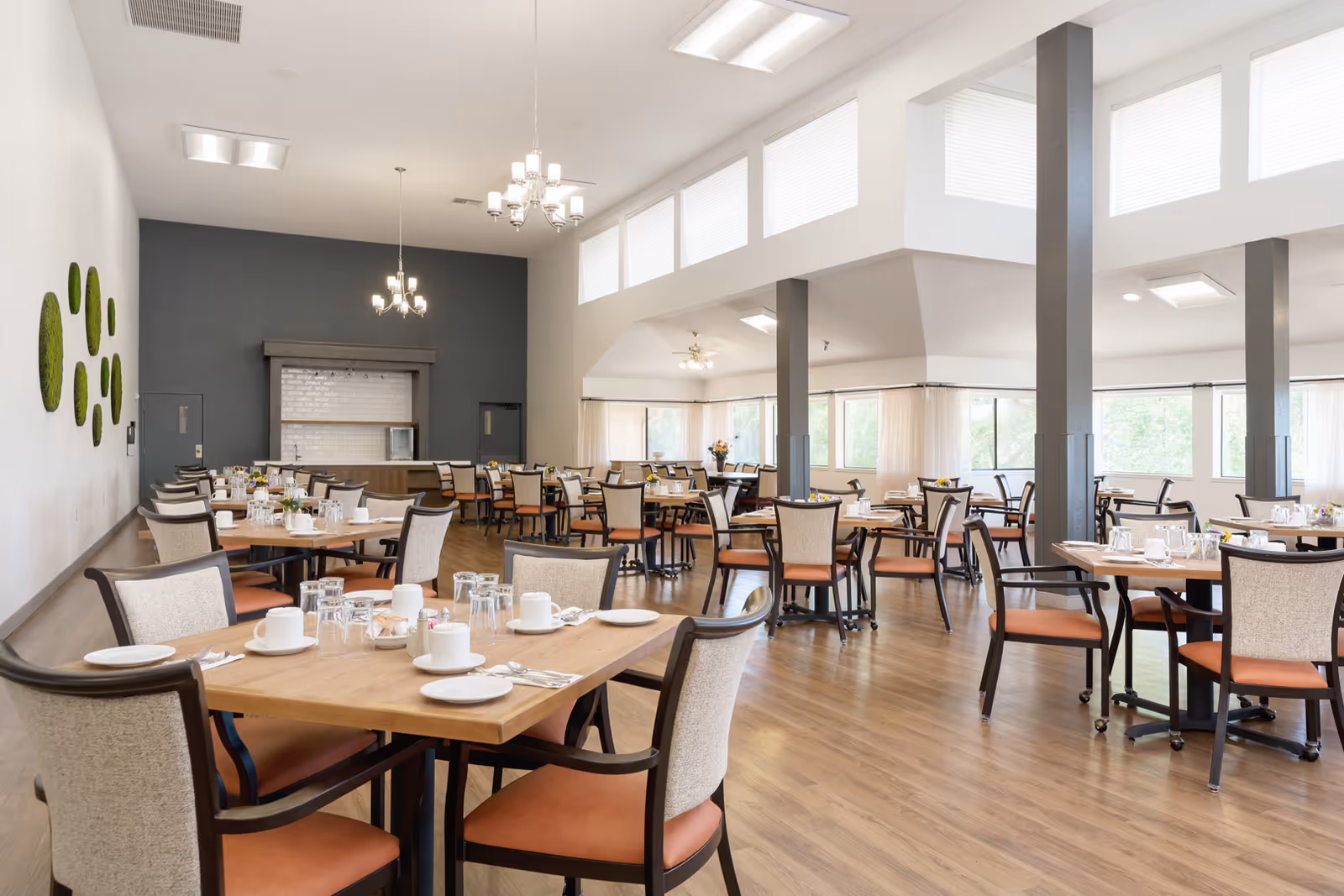 A spacious dining room in a retirement community with multiple wooden tables and cushioned chairs arranged neatly. Each table is set with white plates, cups, glasses, and silverware. The room has large windows allowing natural light to fill the space, light-colored walls with green circular wall decorations, and modern chandeliers hanging from the ceiling. The floor is wooden, and there are several structural columns throughout the room.