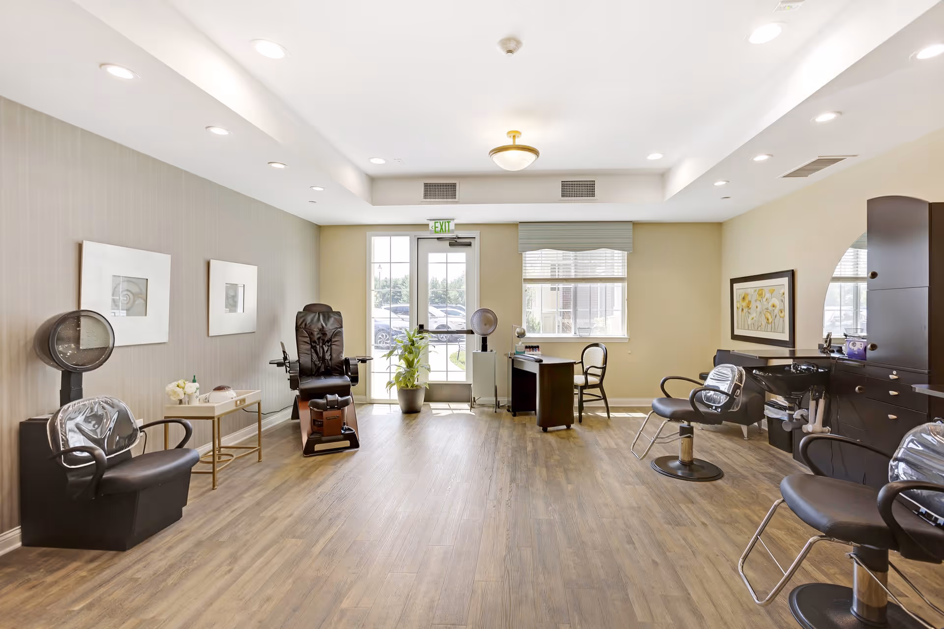 A bright and spacious salon area with wooden flooring, featuring multiple black salon chairs, a hair washing station, a hair dryer, a small desk with a chair, and large windows letting in natural light. The walls are decorated with framed artwork and the room has recessed lighting in the ceiling.