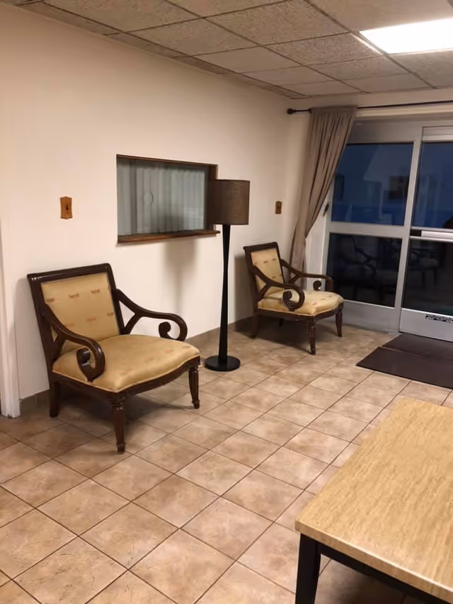 Small lobby-like interior with two upholstered wooden chairs, a floor lamp, tiled floor and a glass entrance door.