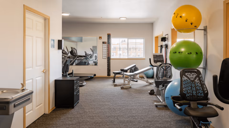 A fitness room with exercise equipment including stationary bikes, a weight bench, and large colorful exercise balls stacked on a rack. There is a window letting in natural light, a water fountain on the left, and a mirror on the far wall reflecting some of the equipment.