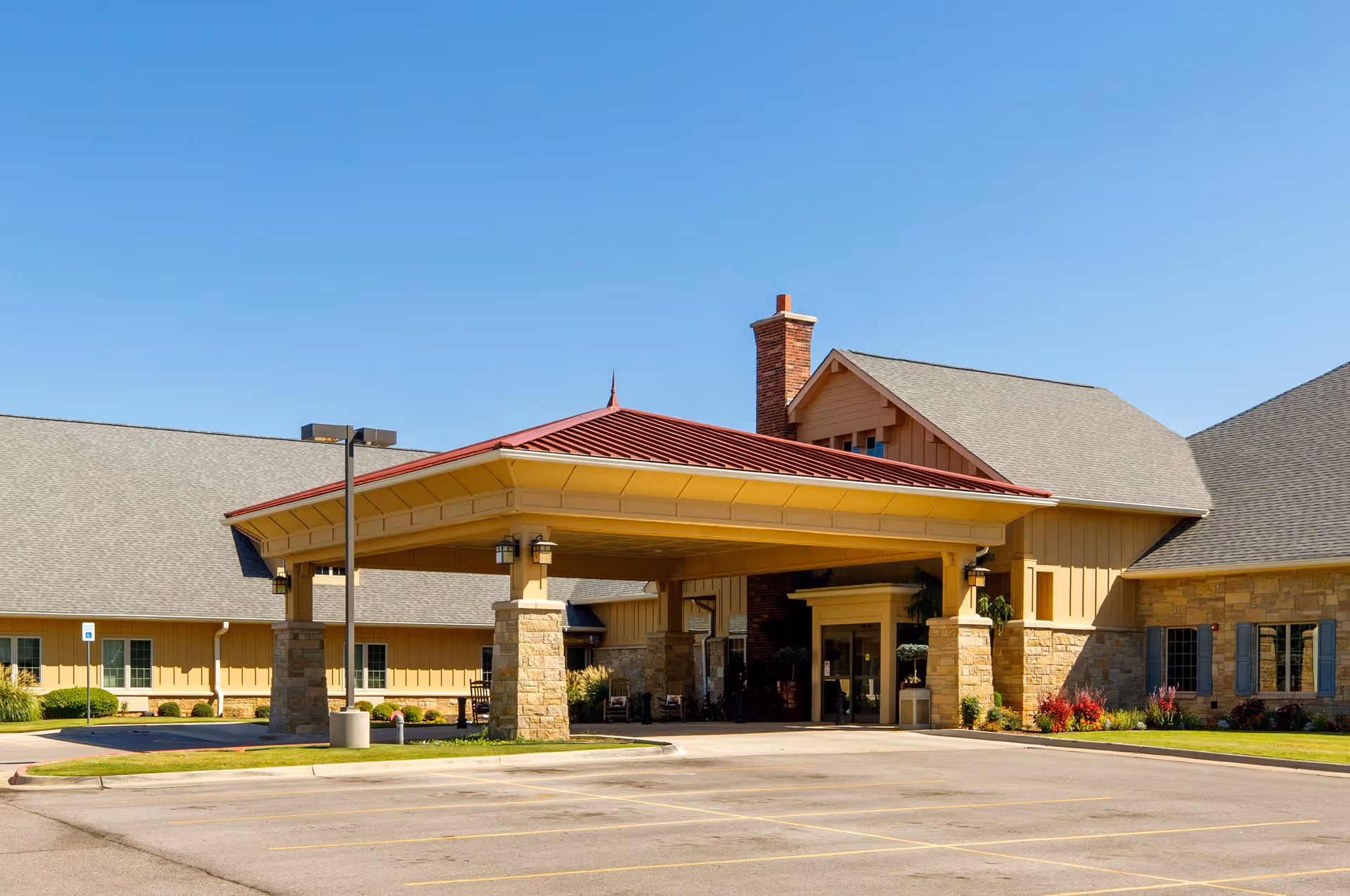 Exterior view of Legend Memory Care at Rivendell showing the main entrance with a covered drop-off area supported by stone pillars, a red roof, and a brick chimney. The building has beige siding and stone accents, with a clear blue sky above and an empty parking lot in front.
