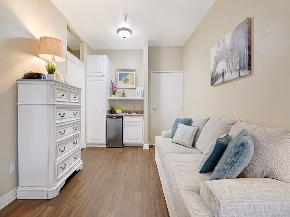 Studio living area with a beige sofa and blue pillows, a white dresser, and a small kitchenette with a mini fridge.