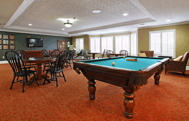 Carpeted community room with a green pool table, round tables and chairs, and seating near large windows.