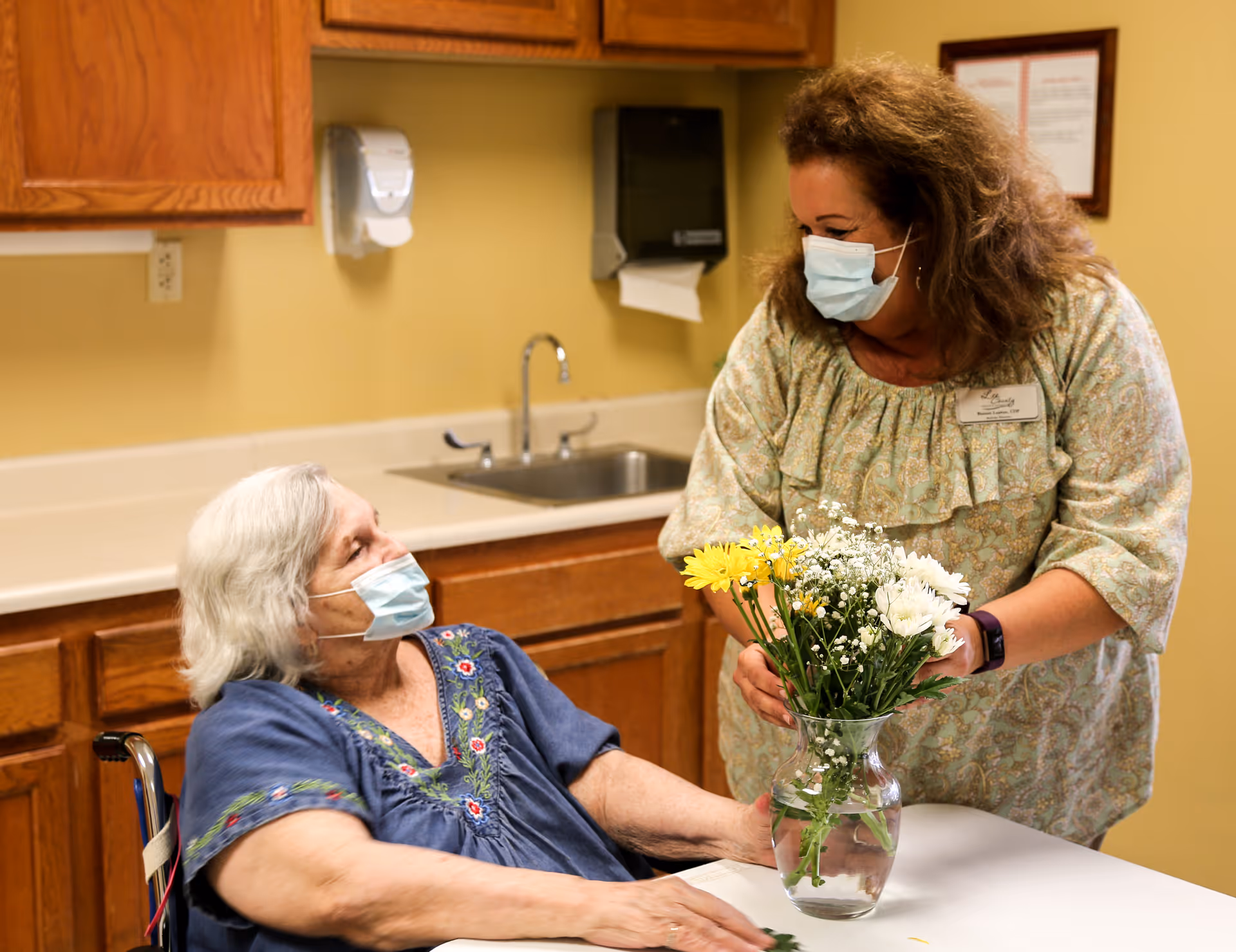 An elderly woman in a wheelchair wearing a blue embroidered top and a face mask is sitting at a table in a room with wooden cabinets and a sink. A woman wearing a patterned blouse and a face mask is arranging a bouquet of yellow and white flowers in a glass vase on the table, smiling at the elderly woman.
