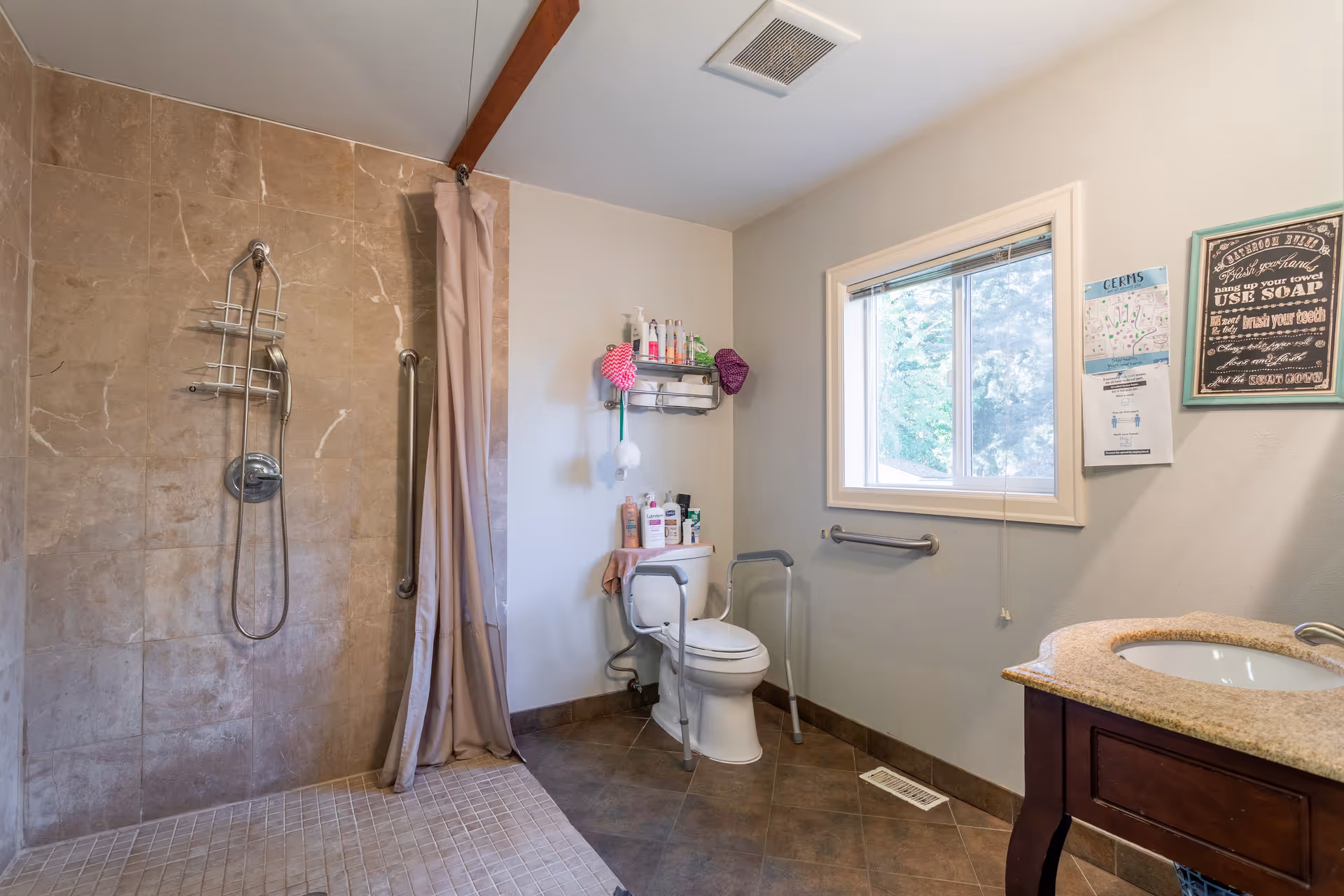 A bathroom with a walk-in shower featuring a handheld showerhead and a beige curtain. There is a toilet with safety rails around it and various toiletries on a shelf above. A window with a view of greenery outside is on the wall next to the toilet. A sink with a granite countertop and wooden cabinet is visible on the right side. The walls are light-colored and the floor is tiled.