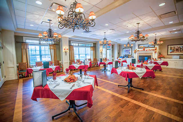 A spacious dining room with multiple tables covered in red tablecloths and white overlays, each set with glassware, silverware, and menus. The room features large windows with curtains, elegant chandeliers hanging from the ceiling, and a fireplace decorated with autumn-themed items.