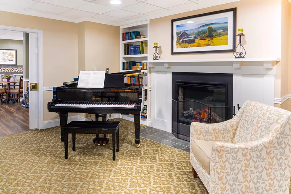 A cozy interior room featuring a black grand piano with sheet music on the stand, a patterned beige armchair, and a white fireplace with a fire burning inside. Above the fireplace is a framed landscape painting, and there are two green candles on decorative holders on the mantel. A bookshelf filled with books is visible behind the piano, and an adjoining room with a dining table and chairs can be seen through a doorway.