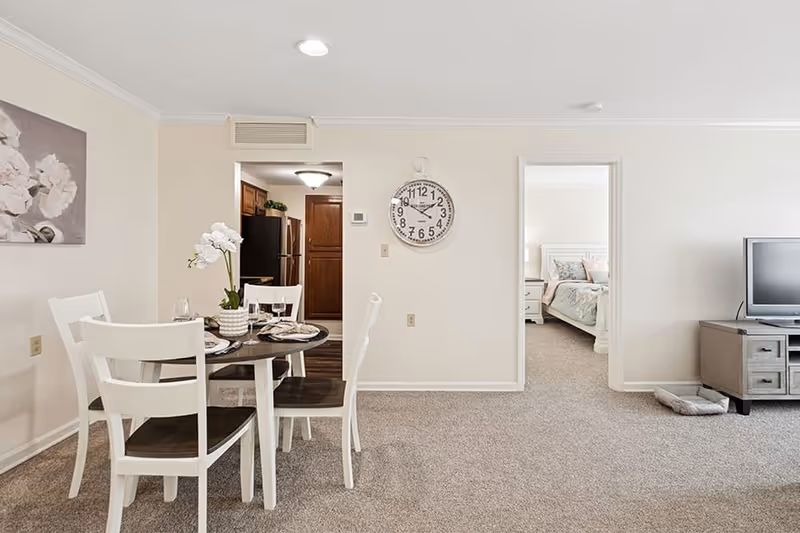 Interior view of a senior living facility showing a dining area with a round table set for four, white chairs, and a flower centerpiece. In the background, there is a kitchen with wooden cabinets and stainless steel appliances. To the right, an open doorway reveals a bedroom with a bed and nightstand. A wall clock is mounted on the wall between the kitchen and bedroom doorways. A TV sits on a wooden stand with a pet bed on the floor nearby.