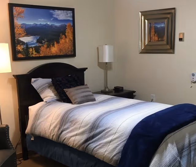 A neatly made single bed with white and blue bedding and multiple pillows in a room. There is a dark wooden headboard, a nightstand with a lamp and a small black item on it, and two framed pictures on the beige walls. A chair is partially visible on the left side.