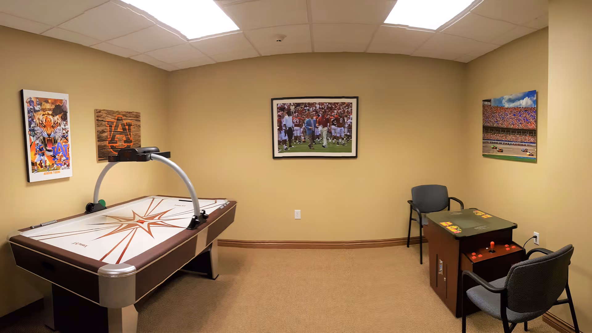 A small game room with beige walls and carpeted floor featuring an air hockey table on the left and a vintage arcade game machine with two chairs on the right. The walls are decorated with framed sports and racing pictures.