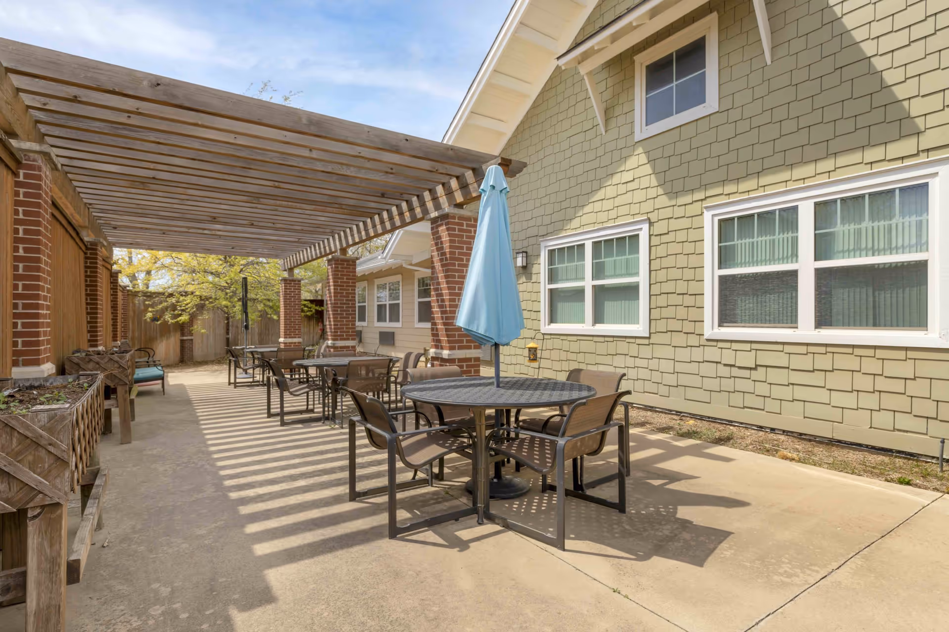 Outdoor patio with round tables and chairs, a blue umbrella, and a wooden pergola beside a green-shingled building.