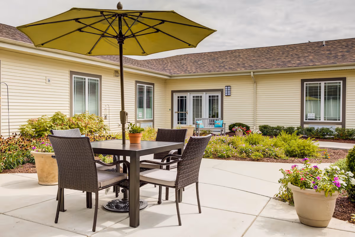 Outdoor patio area at Arbor Terrace Naperville featuring a square table with four wicker chairs and a large green umbrella. The patio is surrounded by flower beds with various plants and flowers, and the building with beige siding and windows is visible in the background.