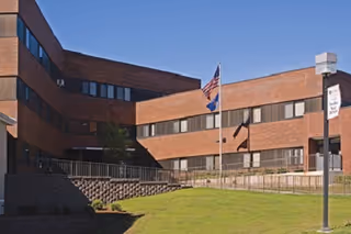 Brick multi-story senior living facility with flagpoles and a manicured lawn under a clear blue sky.