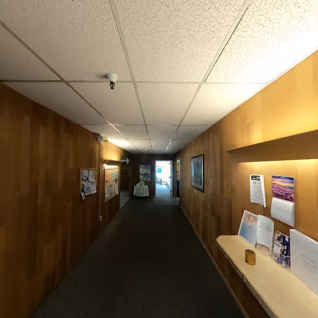 A dimly lit hallway with wood-paneled walls and a drop ceiling. On the right side, there is a built-in shelf with various pamphlets, a calendar, and a small wooden cup. On the left side, there is a bulletin board with pinned papers. At the end of the hallway, there is an open doorway leading to a brighter room with visible furniture.