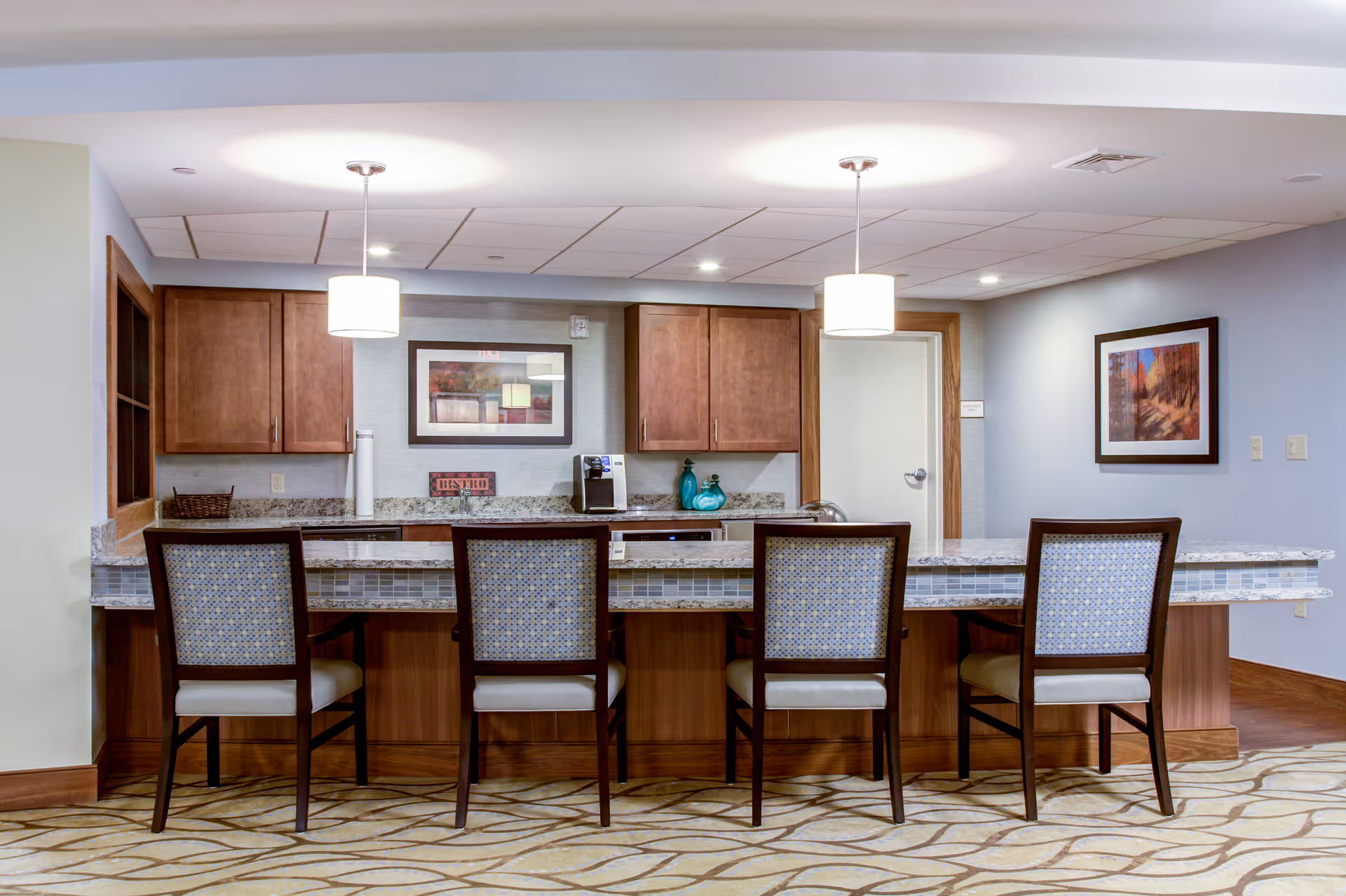Communal kitchen/bar area with four high-backed chairs, granite countertop, pendant lights, and wooden cabinets.