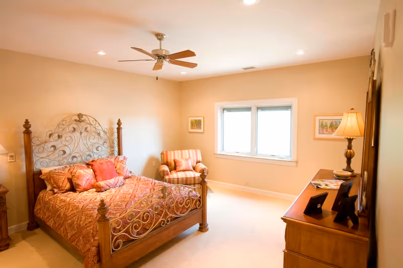 A warmly lit bedroom featuring a wooden bed with an ornate metal headboard and footboard, dressed in a patterned red and gold bedspread with matching pillows. To the left of the bed is a wooden nightstand with a lamp. A striped armchair with a matching pillow is positioned near a window with white trim. On the right side of the room is a wooden dresser with a lamp and framed photos. The walls are painted a soft beige color and the ceiling has a fan with wooden blades.