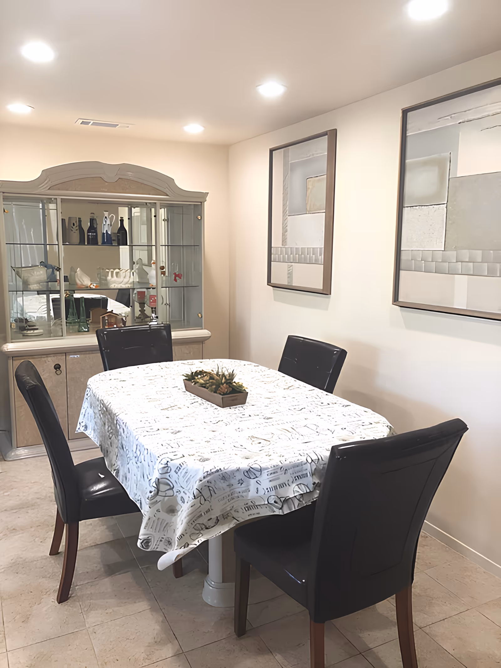 Dining room with a rectangular table covered by a patterned tablecloth, four black chairs, a small centerpiece, framed wall art, and a glass-front china cabinet.