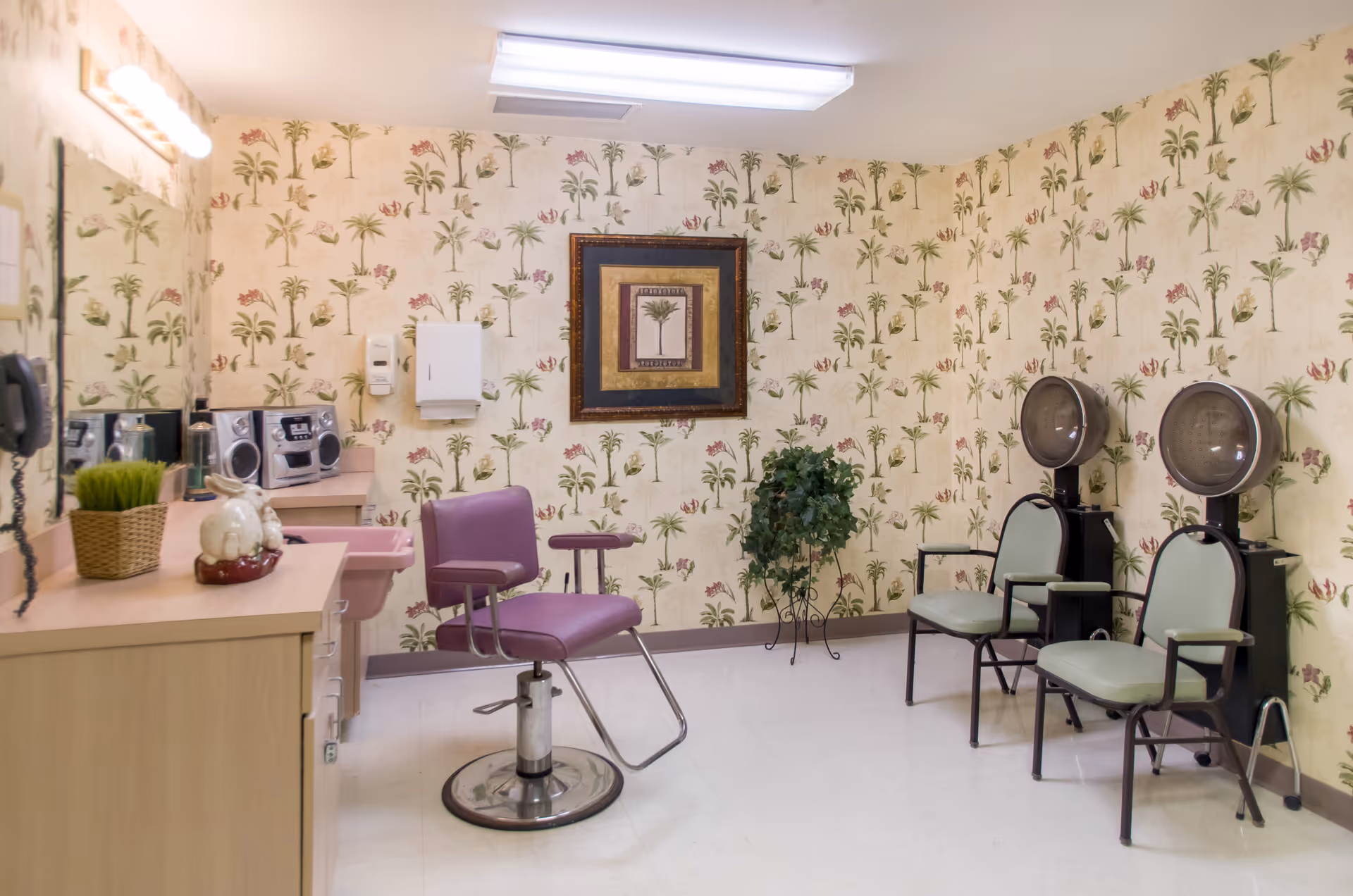 Interior of a salon room with floral wallpaper, featuring a purple salon chair in the center, two green chairs with hair dryers attached on the right, a wooden cabinet with decorative items on the left, and a framed picture on the wall.