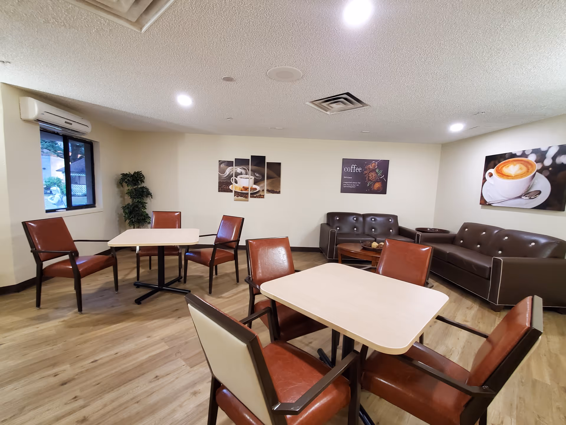 A cozy common area in Diamond Assisted Living & Memory Care featuring two square tables each surrounded by four brown leather chairs. In the background, there are two brown leather sofas with a round wooden coffee table between them. The walls are decorated with three coffee-themed artworks. A window with a view of greenery and an air conditioning unit is visible on the left side. The room has wood flooring and recessed ceiling lights.