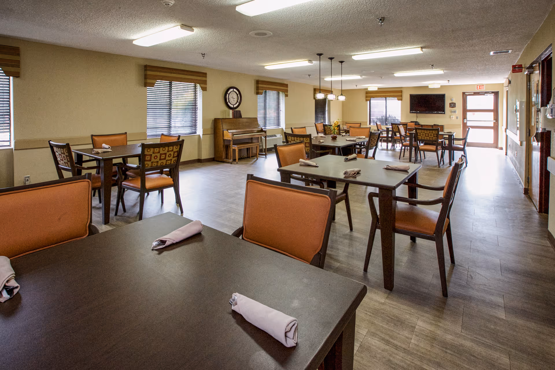 A spacious dining room with multiple dark wood tables and orange cushioned chairs. Each table has neatly rolled napkins placed on it. The room has several windows with blinds, a piano against one wall, a clock above the piano, and a flat-screen TV mounted on the far wall. The floor is covered with wood-like flooring, and the room is well-lit with ceiling lights and hanging pendant lights.