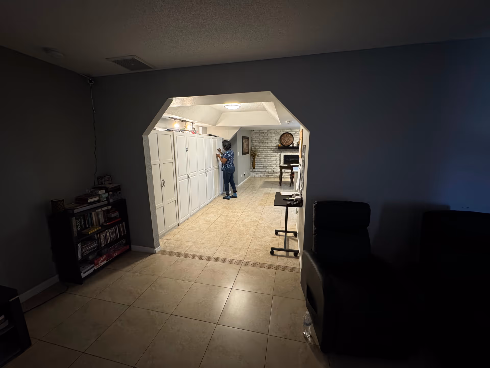 A dimly lit living area opening into a tiled hallway with white storage cabinets and a person standing by them.