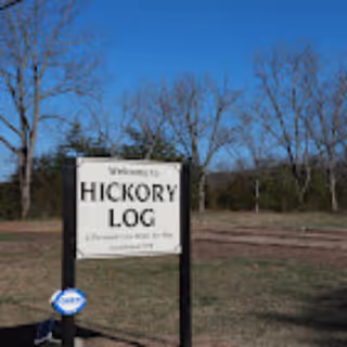 A freestanding sign that reads "Welcome to HICKORY LOG" standing in a grassy field with leafless trees and a clear blue sky.