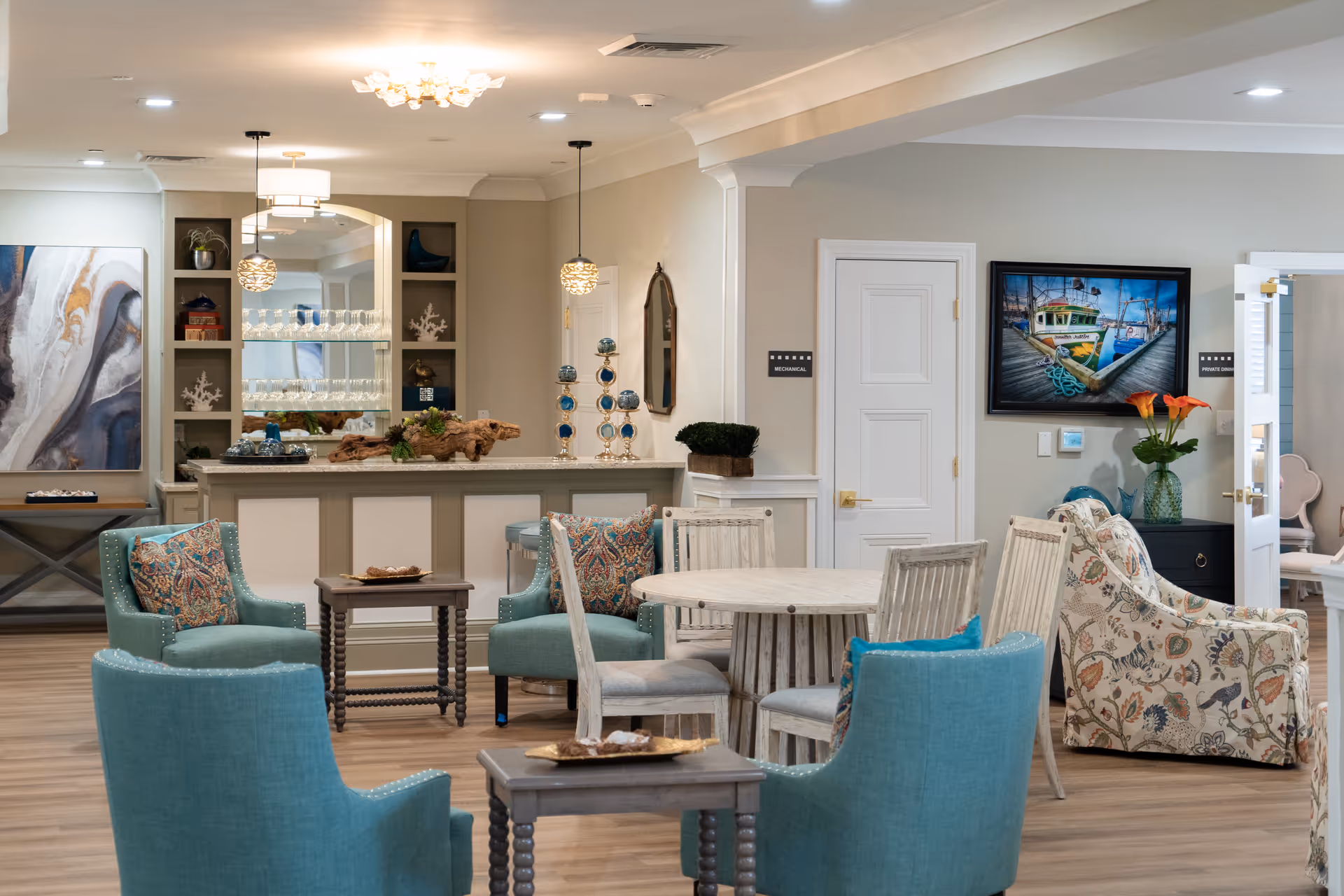 A bright communal lounge with teal armchairs, round wooden tables, patterned sofas, and a service counter/bar in the background.