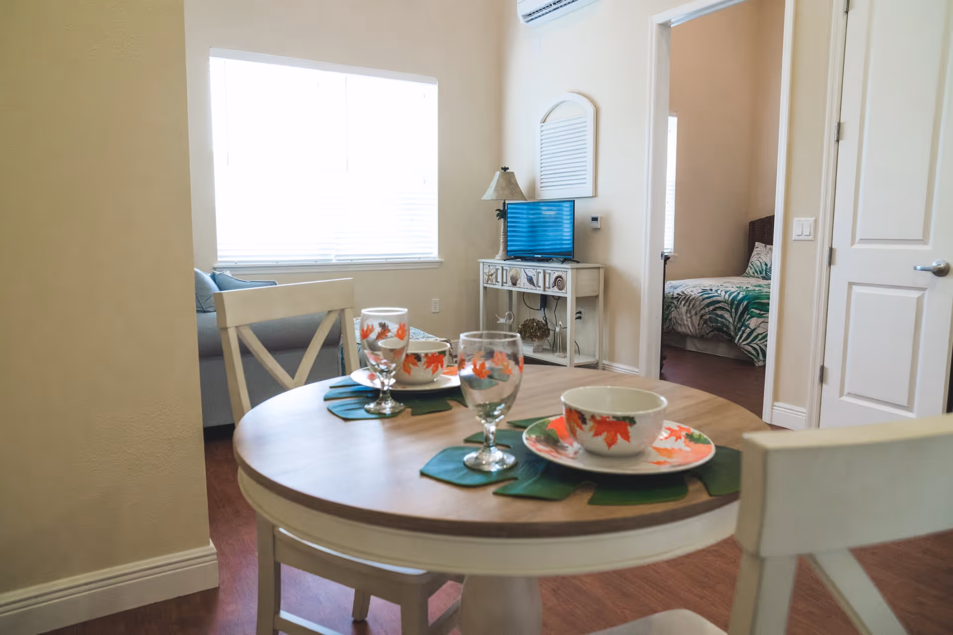 A small dining area with a round wooden table set for two with bowls, plates, and glasses decorated with autumn leaves. In the background, there is a living room with a couch and a TV on a white stand. A bedroom with a bed covered in green and white leaf-patterned bedding is visible through an open door.