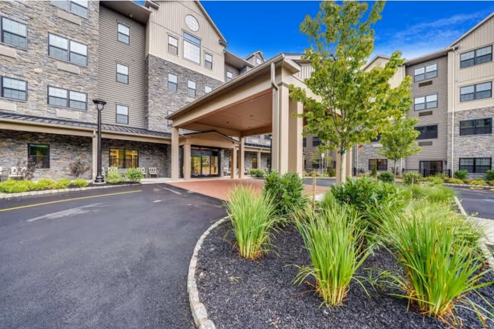 Entrance of a multi-story senior living facility with a covered drop-off area, landscaped greenery including small trees and bushes, and a paved driveway.