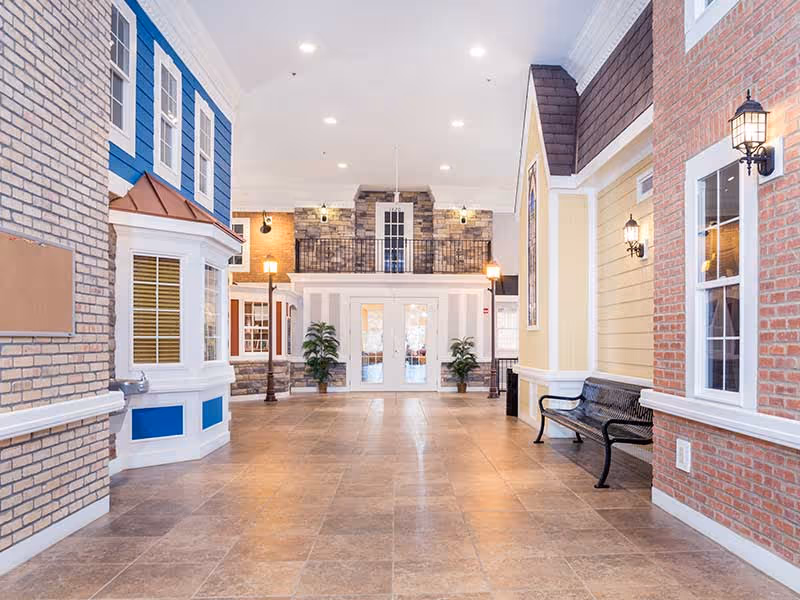 Indoor hallway designed to resemble an outdoor street scene with building facades on either side, including windows, lamps, and a bench. The floor is tiled, and there are potted plants near double glass doors at the end of the hallway.