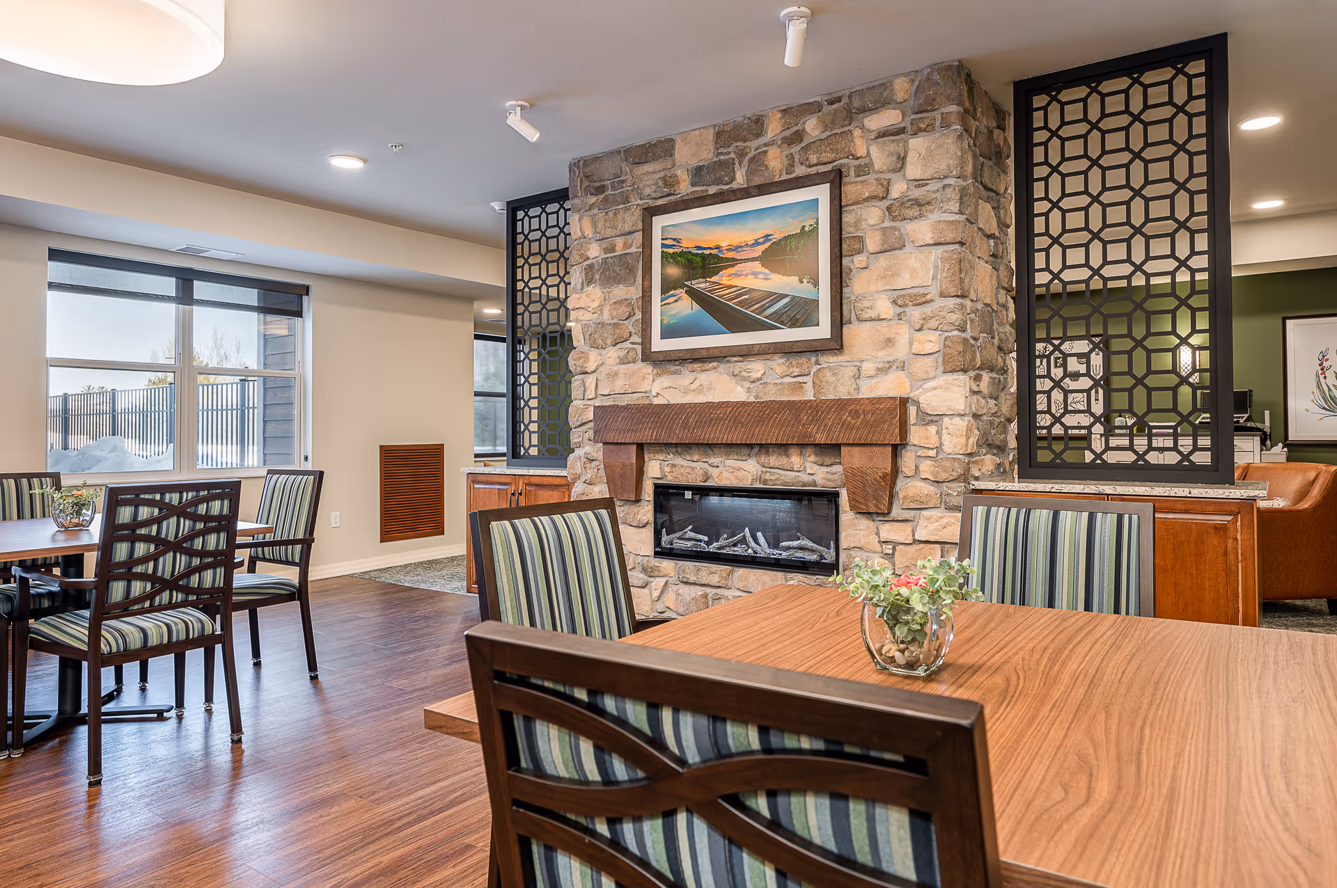 A cozy interior dining area with wooden tables and striped cushioned chairs. A stone fireplace with a wooden mantle is centered on the wall, above which hangs a framed picture of a serene lake scene with a dock. Decorative black lattice panels flank the fireplace. Large windows let in natural light, and the floor is a warm wood tone.
